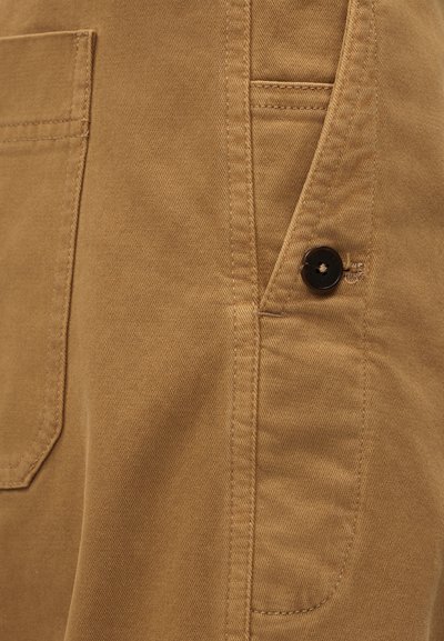 Κοντινό πλάνο ταμπά υφάσματος με λεπτομερείς ραφές, μια μπροστινή τσέπη και ένα μαύρο κουμπί ραμμένο κοντά σε κάθετη ραφή.