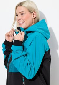 Veste imperméable teal et noire avec capuche, dotée d'une fermeture éclair à l'avant, de poignets élastiques et d'un design coloré. Texture lisse et coupe ajustée.