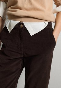 Dunkelbraune Cordhosen mit Seitentaschen, ausgestattet mit einem goldenen Knopfverschluss und einem weißen Hemd, das unter einem beigen Pullover getragen wird.