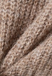 Beige gestrickter Stoff mit einer strukturierten, flauschigen Oberfläche. Das Garn zeigt ein grobes Webmuster, das verschiedene Brauntöne aufweist.
