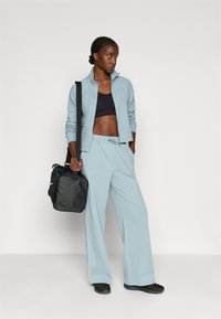 Lyseblå jakke med glidelås og matchende bukser i mykt stoff, kombinert med en svart sportbh, svarte sko og en svart veske.