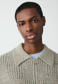 Zbliżenie na osobę z krótkimi włosami, ubraną w beżowy sweter z teksturą na zamek błyskawiczny, noszącą jasnoniebieską koszulę, na jednolitym tle.