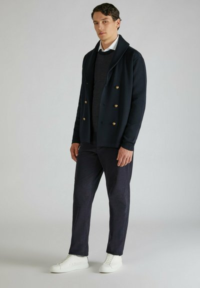 Giacca doppiopetto blu navy con bottoni dorati sopra un maglione di lana scura, abbinata a pantaloni scuri e sneakers bianche. Tessuto dalla texture liscia.