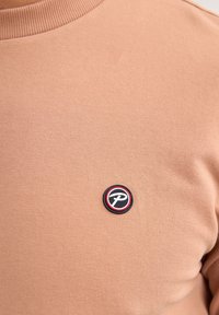Felpa khaki con collo rotondo e texture a costine, con un logo circolare prominente in nero, bianco e rosso sul petto.