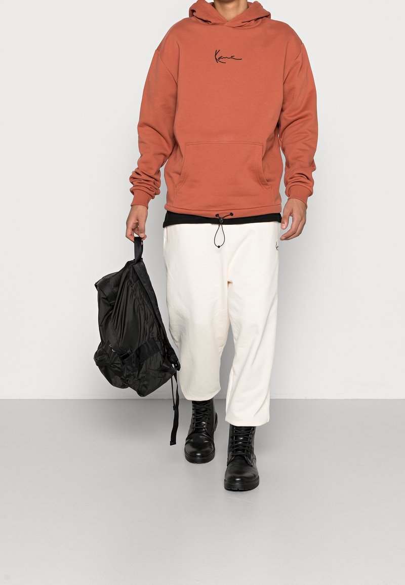 Oranžová mikina s černým logem, bílé kalhoty a černé bojové boty. Drží černý batoh se stahovací šňůrkou. Minimalistický design.