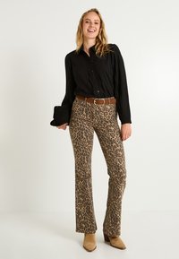 Blusa negra con textura y volantes, combinada con pantalones de campana de estampado leopardo en tonos beige. Cinturón marrón y botas de tacón ankle en marrón claro.