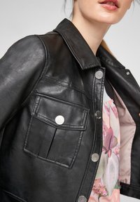 Chaqueta de cuero negra con cuello, con botones a presión plateados y un bolsillo frontal. El interior presenta un diseño floral en tonos rosas.