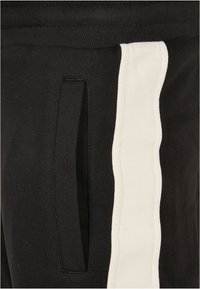 Pantaloni de trening negri cu un buzunar lateral, evidențiați de o dunga albă contrastantă care se desfășoară vertical pe cusătura exterioară.