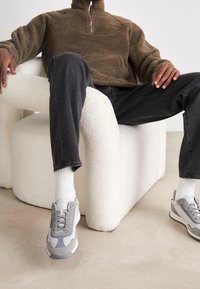 Braune Fleece-Zipjacke, dunkelgraue Jeans, weiße Socken und graue Sneakers mit verschiedenen Texturen, sitzend auf einem weißen skulpturalen Stuhl.