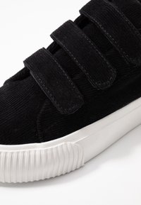 Baskets en velours côtelé noir avec trois bandes velcro et une semelle extérieure épaisse en caoutchouc blanc avec bord avant texturé.