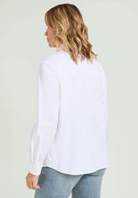 Camicia bianca con bottoni realizzata in tessuto morbido, con maniche lunghe, colletto e orlo arrotondato, indossata con jeans azzurri.