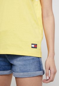 Żółta bawełniana koszulka z białym paskiem, z małą naszywką Tommy Jeans w czerwonym, białym i niebieskim kolorze na dolnym brzegu. Noszona z dżinsowymi szortami.