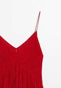Punainen hihaton mekko, jossa on syvä V-mekko, teksturoidut laskokset ja kapeat hihnat. Vyötäröllä on rypytysosa, joka tuo lisää muotoa.