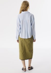 Blusa de manga larga color azul claro con un pliegue en la espalda, combinada con una falda verde olivo hasta la rodilla con detalles de dobladillo sin terminar y sandalias de tacón negras.