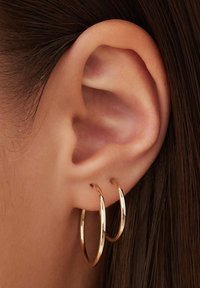 Des boucles d'oreilles en or pendent d'un lobe d'oreille percé, réfléchissant la lumière avec une finition polie. Le design présente deux grands anneaux lisses.