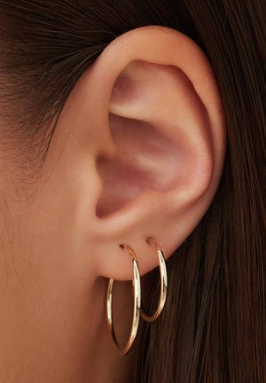 Des boucles d'oreilles en or pendent d'un lobe d'oreille percé, réfléchissant la lumière avec une finition polie. Le design présente deux grands anneaux lisses.