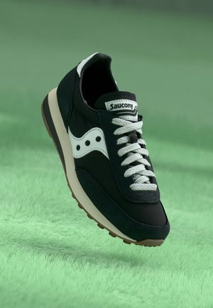 Sort-hvid Saucony løbesko med hvide snørebånd, der svæver over en grøn tekstureret overflade.