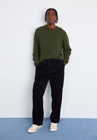Pull en tricot vert olive à col rond, associé à un pantalon en velours côtelé bleu marine et des baskets blanches avec des accents rouges, se tenant devant un fond uni.