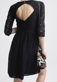 Schwarzes Spitzenkleid mit Dreiviertelärmeln, einem weit schwingenden Rock und einem Schlüssellochausschnitt hinten. Kombiniert mit einer gemusterten Umhängetasche.