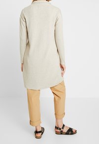 Beige ribbad tunika med långa ärmar, hög-låg fåll, ihop med uppvikta beige byxor och svarta remsandaler mot en ljus bakgrund.