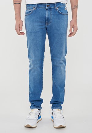 Jeans in denim azzurro chiaro con una vestibilità slim, caratterizzati da cuciture arancioni e un classico design a cinque tasche. Indossati con sneaker bianche.