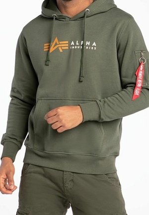 Mann trägt olivgrünen Alpha Industries Hoodie mit Fronttasche und rotem "Remove Before Flight" Anhänger an der Reißverschlusstasche am linken Ärmel.