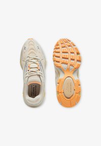 Zapatillas deportivas color beige claro con parte superior de malla, detalles en plateado y suela naranja. Cuentan con goma texturizada para agarre y cuello acolchado.