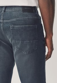 Gros plan d'une personne portant un jean denim bleu foncé avec une poche arrière visible et une étiquette en denim sur la ceinture.