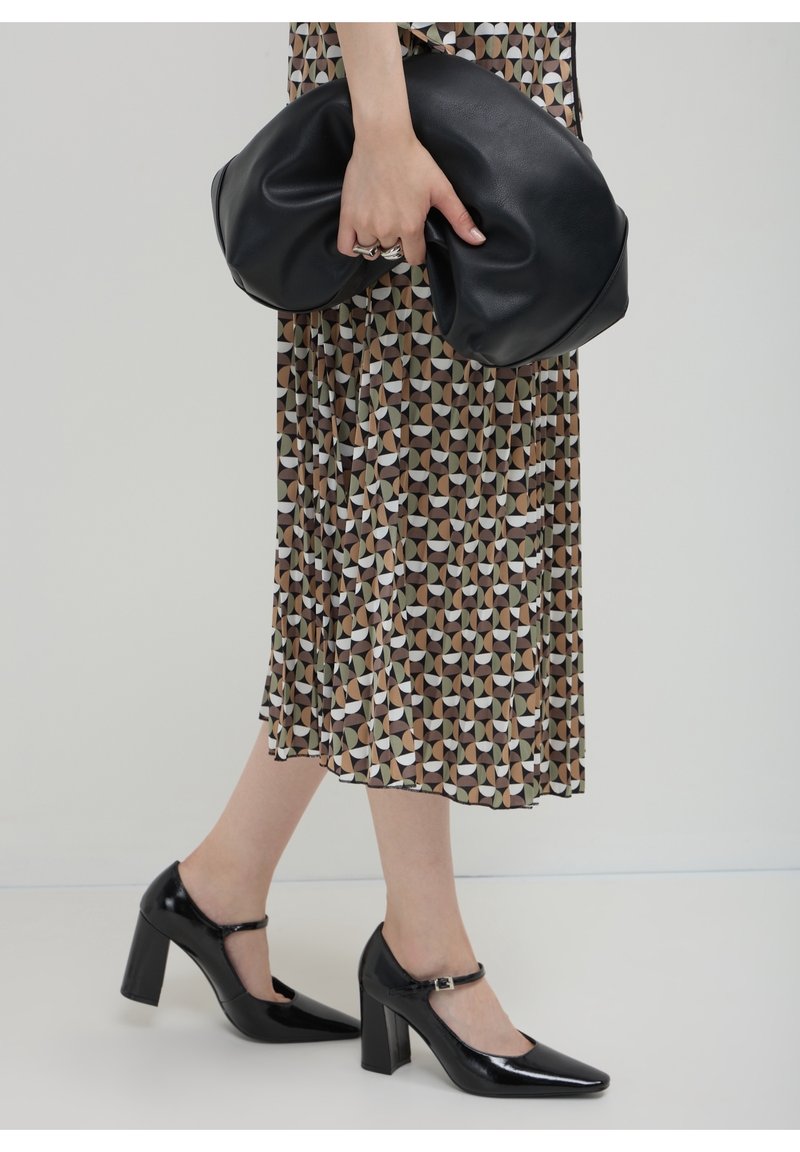 Personne portant une jupe midi à motifs géométriques, des escarpins Mary Jane en cuir verni noir, tenant une grande pochette souple noire dans une main.