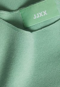 Tkanina u boji mente s glatkom teksturom, na kojoj se vidi oznaka s natpisom "JJXX." Materijal prikazuje fini rebrasti uzorak.