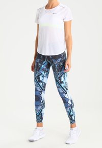 Maglietta Nike bianca a maniche corte in mesh con striscia neon, abbinata a leggings colorati con motivo geometrico in blu e nero.