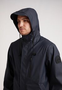 Veste imperméable navy avec capuche, dotée d'une poche zippée, de cordons de serrage réglables et d'un écusson sur la manche gauche. Texture lisse.
