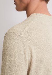 Primo piano della spalla e del collo di una persona che indossa un maglione a maglia beige con una texture, visto da dietro.
