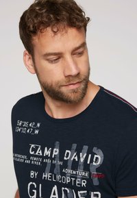 Camiseta azul marino de tejido suave con cuello redondo, texto gráfico y acentos a rayas rojas y blancas en los hombros.