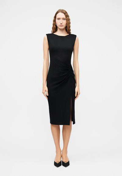 Femme aux cheveux ondulés portant une robe noire midi sans manches, ajustée, avec fronces sur le côté et une fente haute à l'avant, associée à des escarpins noirs pointus.