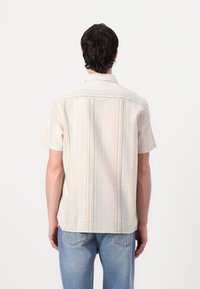 Kortermet skjorte i lys beige stoff med vertikale striper i blått og gult. Har standard krage og bakstykke-detalj.