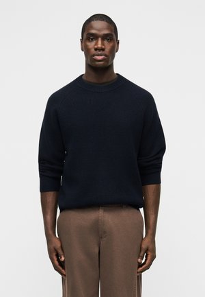 Muž stojaci čelom vpred, oblečený v čiernom rebrovanom svetri a hnedom nohaviciach, na jednoduchom svetlom pozadí.