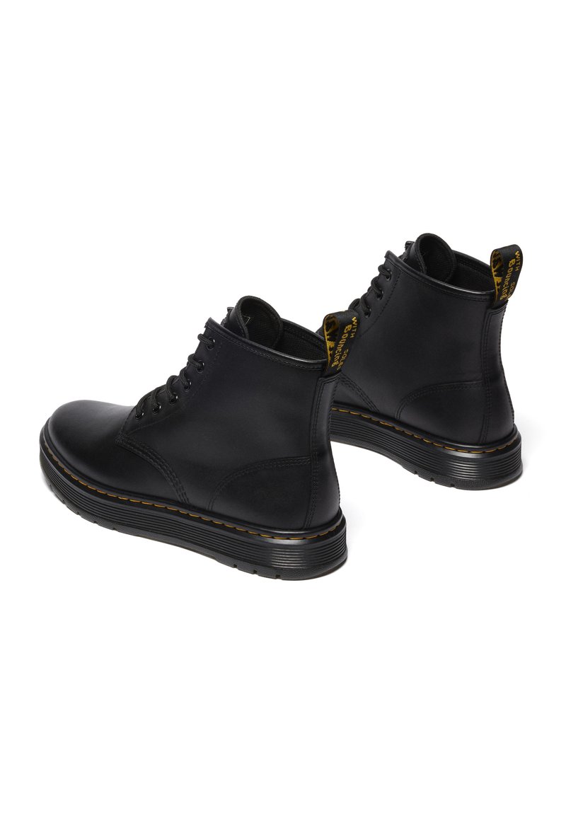 Bottines en cuir noir avec bout rond, lacets à six œillets et tirette texturée jaune. Elles possèdent une semelle noire épaisse et texturée avec des coutures jaunes.