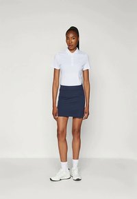 Biela polo košeľa s krátkymi rukávmi, námornícky športový sukňa a biele tenisky. Hladká tkanina, priliehavý strih, klasický golier a detaily s gombíkmi.