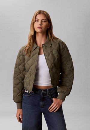 Frau trägt eine olivgrüne gesteppte kurze Jacke über einem weißen Crop-Top und dunkelblauen Jeans, steht vor einem schlichten grauen Hintergrund.