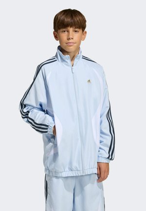 Niño con chándal Adidas azul claro con rayas negras en las mangas y los pantalones, de pie con una mano en el bolsillo contra un fondo liso.