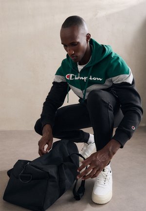 Sudadera deportiva en verde, gris y negro con el logo de Champion; bolsa de deporte negra; zapatillas blancas; texturas y contrastes sutiles.