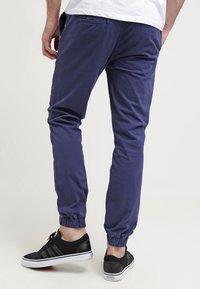 Man in navy blauwe joggerbroek en zwarte Adidas sneakers, staande tegen een effen achtergrond, van achteren weergegeven.