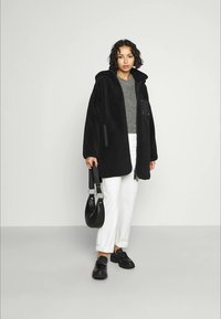 Woman wearing a black hooded coat, gray sweater, white pants, black shoes, holding a black handbag, standing against a plain light background.