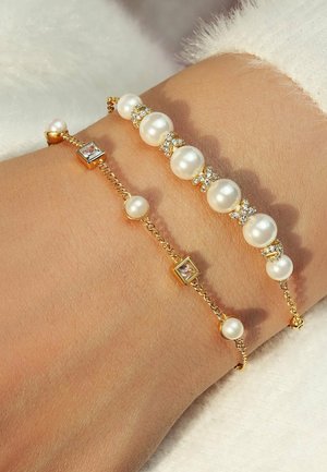 Zwei goldene Armbänder an einem Handgelenk: eines mit großen weißen Perlen und klaren Kristallen, das andere mit kleinen Perlen und quadratischen Akzenten.