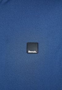 Blue fabric shirt featuring a square black patch with "Bench." printed in white. Smooth texture, simple design, and no additional accents.