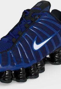 Μπλε και μαύρο αθλητικό παπούτσι Nike Shox με λευκό λογότυπο Swoosh, επάνω μέρος από πλέγμα και χαρακτηριστικά μαύρα ελατηριωτά πέλματα.
