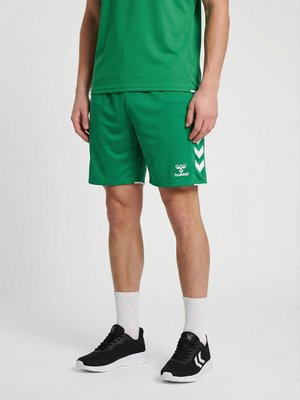 Mand iført grønne sportsshorts og matchende skjorte med sorte og hvide sneakers, der står på en ensfarvet baggrund.