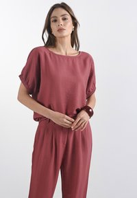 Burgundská texturovaná blůza s širokými rukávy, doplněná o odpovídající kalhoty. Nápadný lesklý náramek na zápěstí přidává kontrast. Jednoduchý, uvolněný design.