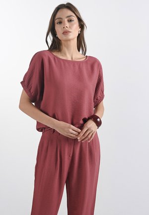 Burgundská texturovaná blůza s širokými rukávy, doplněná o odpovídající kalhoty. Nápadný lesklý náramek na zápěstí přidává kontrast. Jednoduchý, uvolněný design.